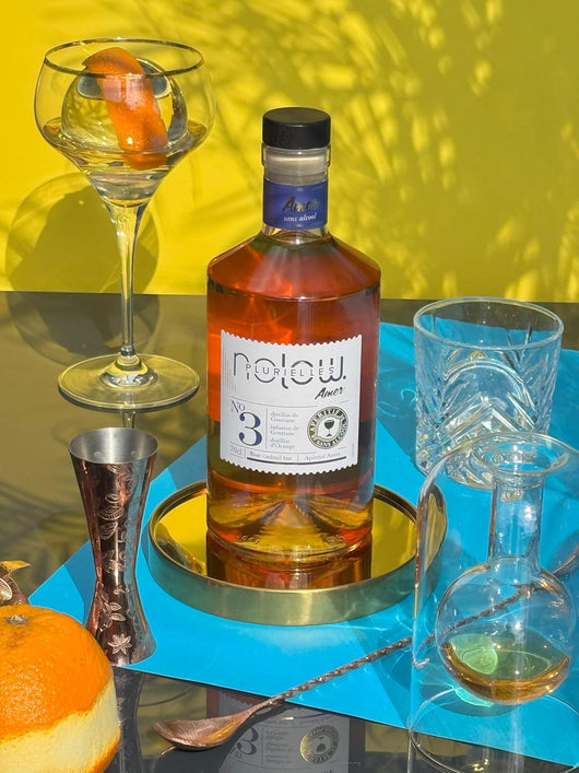 Bottiglie di NOLOW N°3 Distillat Amer analcolico con bicchieri da cocktail e arancia su una tavola colorata.
