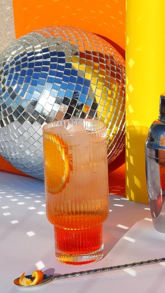 Cocktail in vetro strutturato con contorno arancione, palla da discoteca e sfondo luminoso, ideale per vivaci ambientazioni per feste.